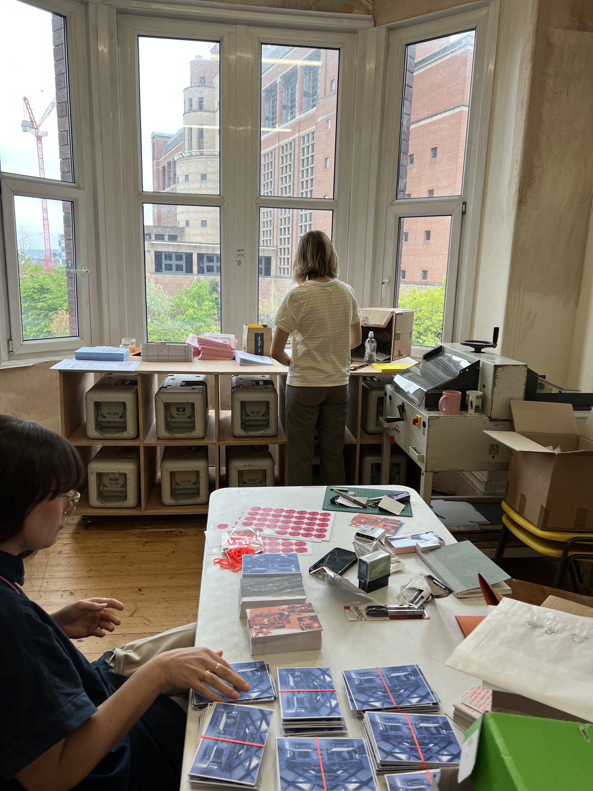 Picture of Daisy from the back arranging piles of printed paper. She is standing in front of several large ink canisters. To the front of the photo is a table with piles of beautiful, colourful paper on top. Kate West, Guild Senior Producer, is sat at the table on the left.
