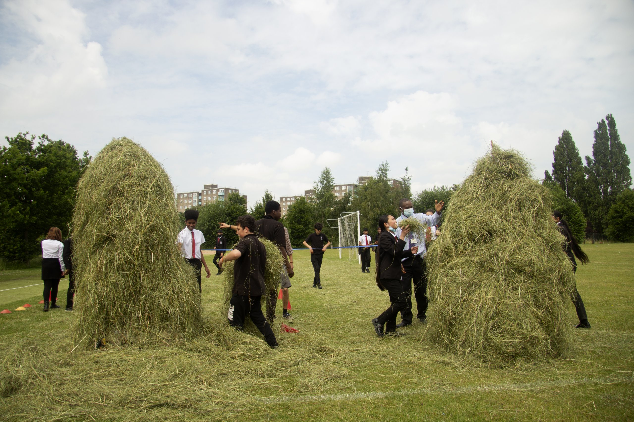 Children from the local community haymaking with Luke from Meanwood Valley Farm. There are two bales of hay either side of the photo. They are tall and surrounded by children.